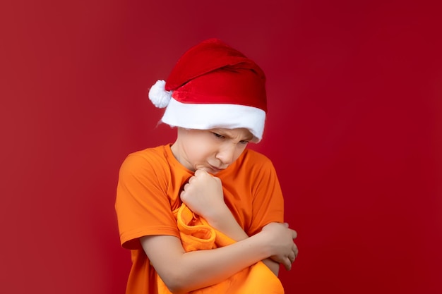 Un niño con una camisa amarilla se encuentra sobre un fondo rojo con un gorro de Papá Noel sosteniendo una bolsa de regalo en sus manos