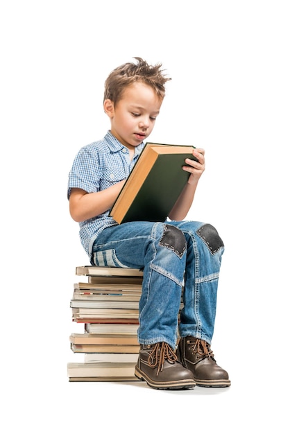 Niño con camisa azul leyendo un libro sentado en una pila de libros sobre un fondo blanco aislado