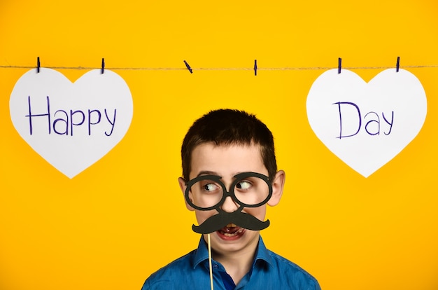 Foto un niño con una camisa azul sobre un fondo amarillo con un corazón y una inscripción, un día festivo está torcido y viste gafas y bigote