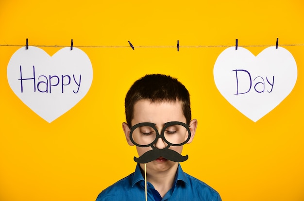 Foto un niño con una camisa azul sobre un fondo amarillo con un corazón y una inscripción, un día festivo está torcido y viste gafas y bigote