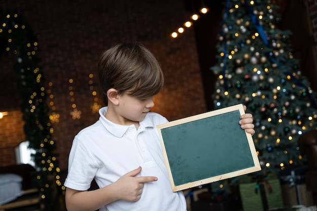 Un niño con una camisa blanca se encuentra junto al árbol de Navidad y sostiene un tablero con un espacio en blanco para el texto. Concepto de vacaciones de Navidad