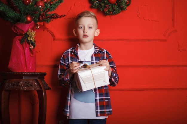 Foto niño con una camisa a cuadros con un regalo en sus manos se encuentra cerca de la pared roja en nochebuena