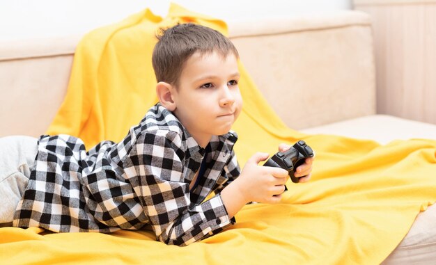 El niño con camisa a cuadros tirado en el sofá con un joystick negro en las manos jugando al videojuego Jugar videojuegos en el concepto de casa