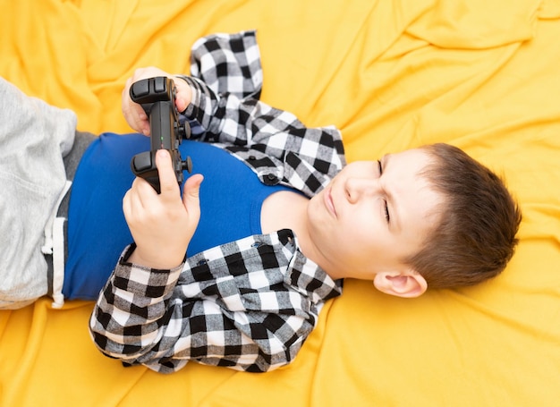El niño con camisa a cuadros tirado en el sofá con un joystick negro en las manos jugando al videojuego Jugar videojuegos en el concepto de casa
