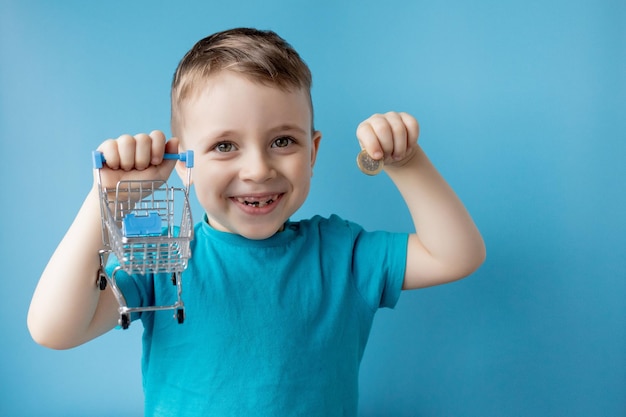 Niño en camiseta azul sostiene carrito de compras y moneda sobre fondo azul Concepto de compras y venta