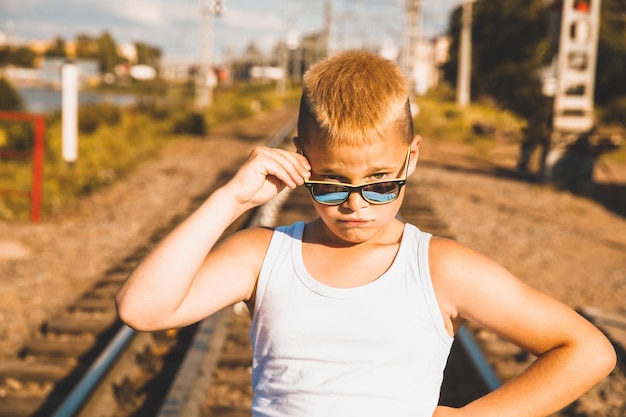 Un niño con una camiseta blanca y gafas negras está parado en el ferrocarril. tono marrón