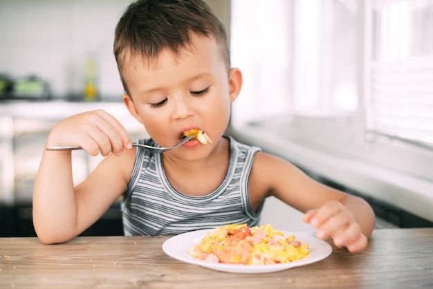 Un niño con una camiseta en la cocina comiendo una tortilla con salchichas y tomates con un tenedor