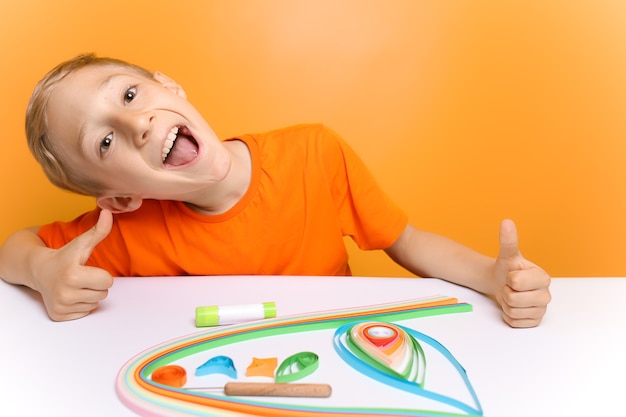 Un niño con una camiseta naranja se ríe de la cámara antes de realizar la torsión de finas tiras de papel en la técnica de quilling.