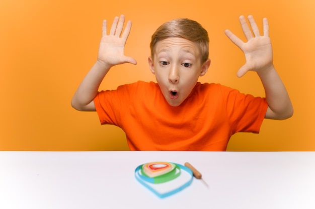 Un niño con una camiseta naranja se sienta en una mesa blanca y emocionalmente levanta las manos y mira el trabajo realizado con tiras de papel delgadas en la técnica de quilling.