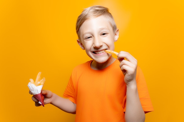 Un niño con una camiseta naranja sostiene una bolsa de papas fritas frente a él y muerde un trozo de papas sonriendo