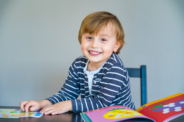 Niño con camiseta a rayas sentado en la mesa, aprendiendo en casa durante la cuarentena por coronavirus. Niño jugando con pegatinas con forma.