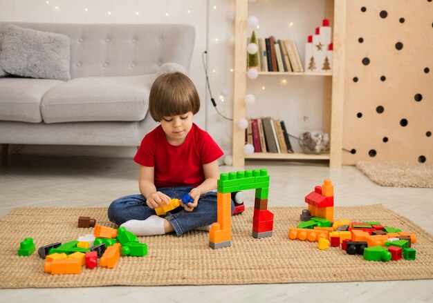 Niño con una camiseta roja y jeans juega con un kit de construcción de colores en el piso junto al sofá de la habitación