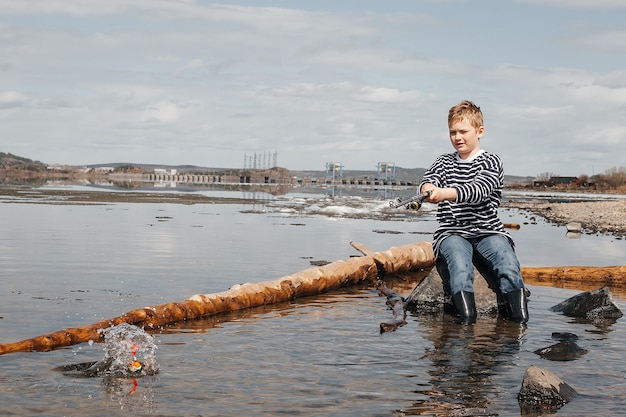 Un niño con una caña de pescar está pescando, sentado en la orilla del río. Un chico guapo con un chaleco a rayas se sienta con una caña de pescar en sus manos