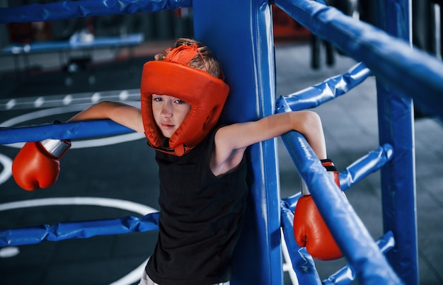 Niño cansado en equipo de protección apoyado en los nudos del ring de boxeo.