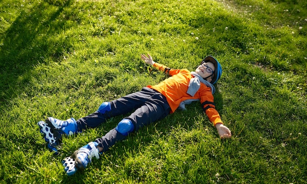 Niño cansado feliz con patines y casco descansa sobre hierba verde