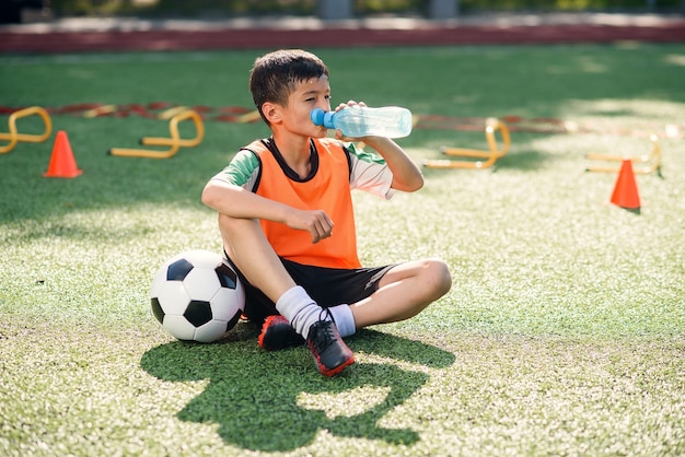 Niño cansado en uniforme de fútbol bebe agua de una botella de plástico después del entrenamiento