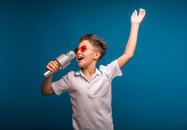 Un niño canta en un micrófono sobre un fondo azul. Un niño guapo con una camiseta blanca y pantalones cortos se para sobre un fondo azul y canta emocionalmente en el micrófono.