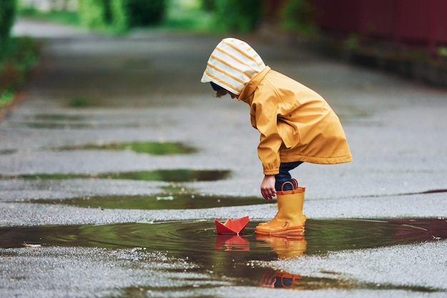 Niño con capa impermeable amarilla y botas jugando al aire libre después de la lluvia