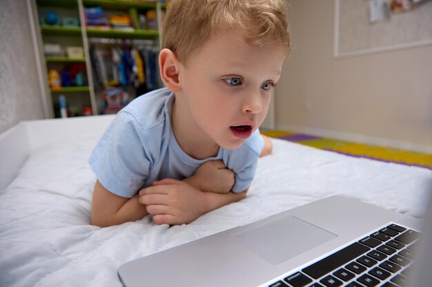 Niño con una cara muy sorprendida acostado en una cama frente a una pantalla de computadora portátil