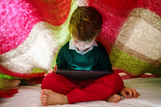 Niño en casa debajo de la manta con tableta