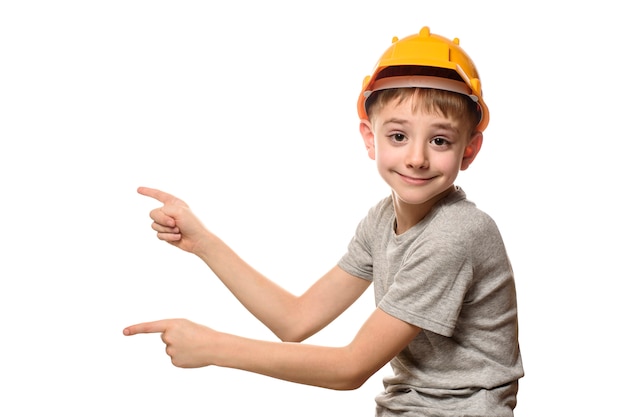 Niño en casco de construcción naranja muestra los dedos índices a un lado. Retrato. Aislar en la pared blanca.