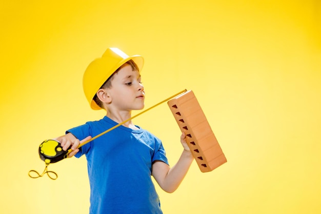 Un niño con casco de construcción sostiene un ladrillo en sus manos y lo mide con una cinta métrica que se queda en el estudio con fondo amarillo