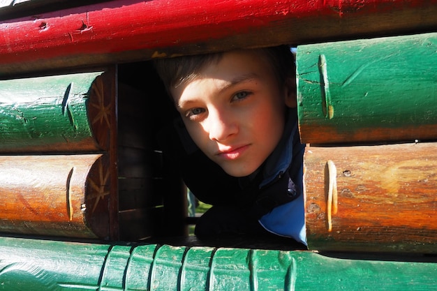 Un niño caucásico de 10 años con cabello rubio y ojos grises mira por la ventana de una casa de madera en un parque infantil Peekaboo El niño se escondió Cara bonita