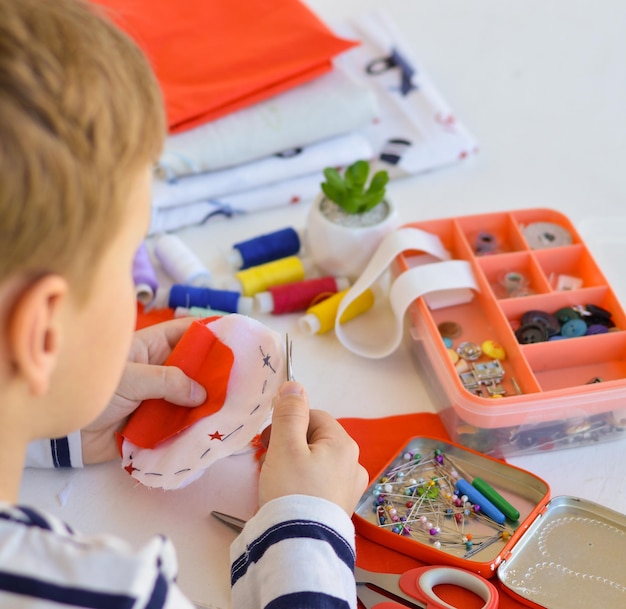 Un niño caucásico de 9 años está aprendiendo a coser con las manos un regalo  casero para mamá para unas vacaciones