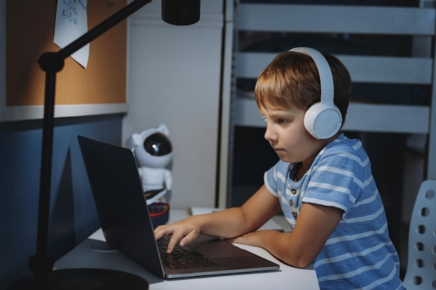 Un niño caucásico con auriculares inalámbricos estudiando en casa escribiendo en una computadora portátil