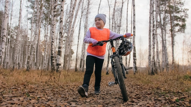 Un niño caucásico camina con bicicleta en el parque de otoño Niña pequeña caminando en ciclo naranja negro