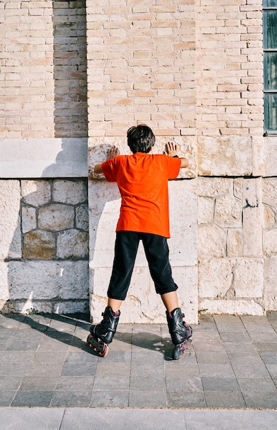 Niño caucásico por detrás jugando con patines y pegado a la pared del parque.