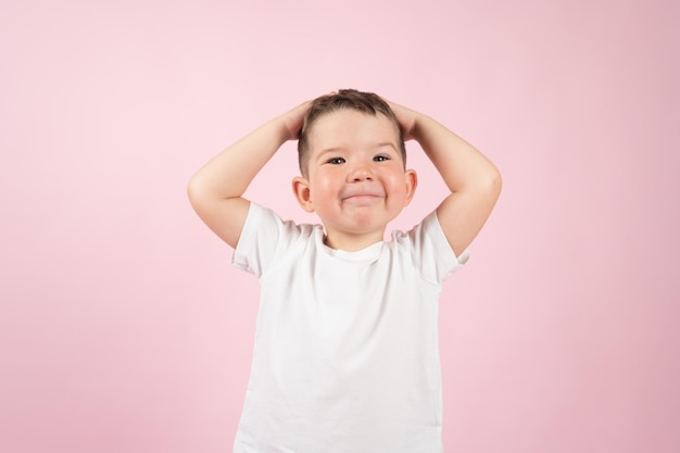 Niño caucásico emocional haciendo muecas y llevándose las manos a la cabeza superficie rosa Foto de alta calidad