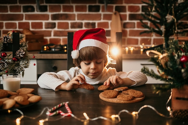 Un niño caucásico está sentado en la mesa de la cocina con un suéter blanco y un sombrero rojo de Navidad con galletas de avena