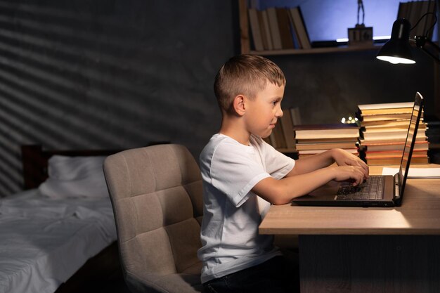 Niño caucásico estudiando por la noche en casa usando una computadora portátil sentado en la mesa niño de escuela aprendiendo aprendizaje electrónico en línea