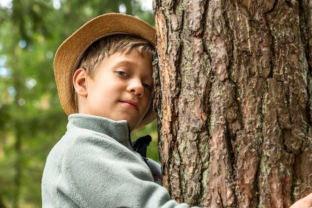 Un niño caucásico feliz en un sombrero abraza un árbol paseos al aire libre