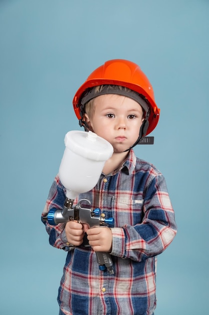 Un niño caucásico feliz con sombrero duro sosteniendo una pistola de pulverización eléctrica, un pintor o un decorador.