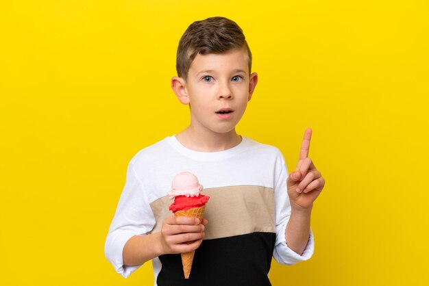 Un niño caucásico con un helado de cucurucho aislado en un fondo amarillo pensando en una idea apuntando con el dedo hacia arriba