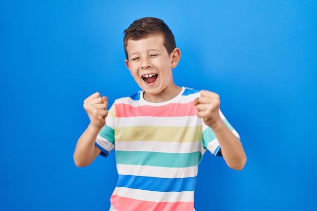 Foto niño caucásico joven parado sobre fondo azul muy feliz y emocionado haciendo gesto ganador con los brazos levantados sonriendo y gritando por el concepto de celebración de éxito