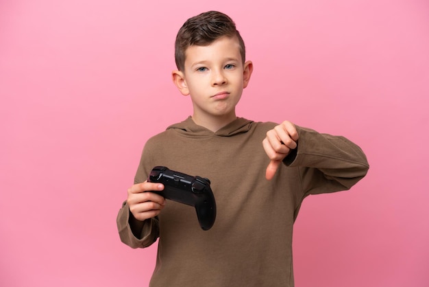 Un niño caucásico jugando con un controlador de videojuegos aislado en un fondo rosa mostrando el pulgar hacia abajo con expresión negativa