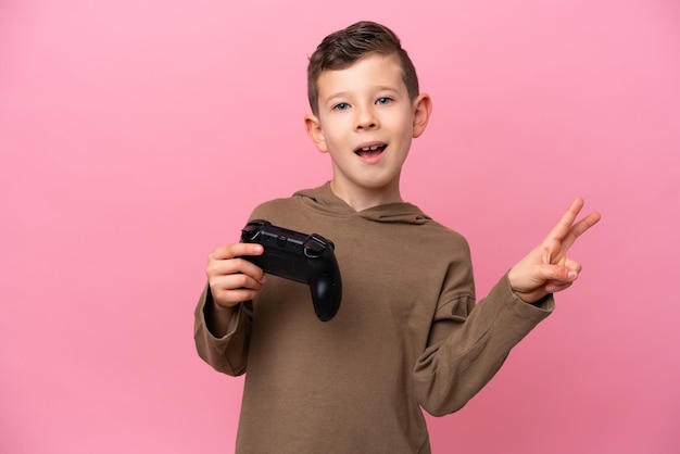 Un niño caucásico jugando con un controlador de videojuegos aislado en un fondo rosa sonriendo y mostrando el signo de la victoria