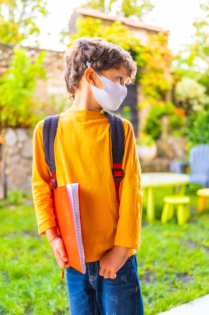 Niño caucásico con mascarilla listo para volver a la escuela. Nueva normalidad, distancia social, pandemia de coronavirus, covid-19. Camiseta naranja, mochila y bloc de notas en mano