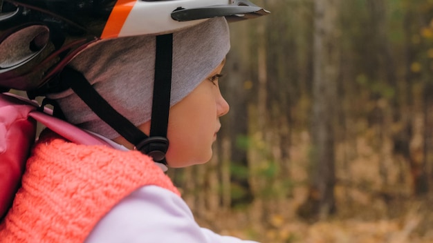 Foto un niño caucásico monta en bicicleta por la carretera en el parque de otoño niña montando un ciclo naranja negro en el bosque niño va a hacer deportes en bicicleta paseo en movimiento de motorista con mochila y casco cola dura de bicicleta de montaña