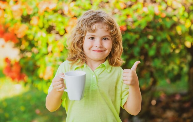 Niño caucásico optimista agua potable sostenga la taza en las manos y sonría aislado sobre la naturaleza verde backg