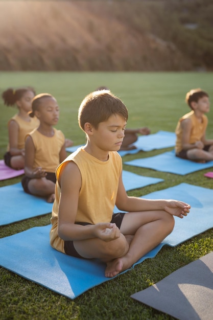 Foto niño caucásico practicando yoga y meditando sentado en una alfombra de yoga en el jardín de la escuela. concepto de escuela y educación
