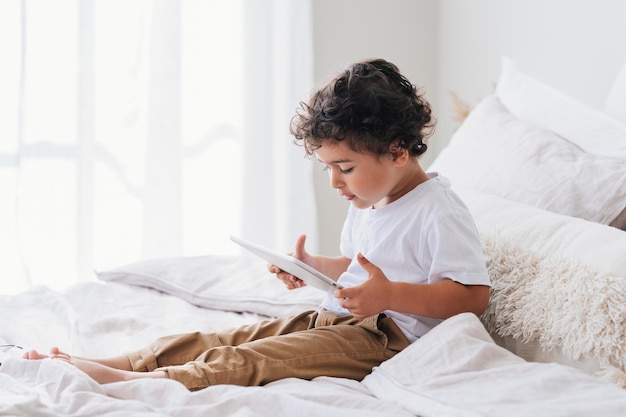 Un niño caucásico rizado se sienta en la cama en casa usando una tableta por la mañana con un espacio vacío en el fondo