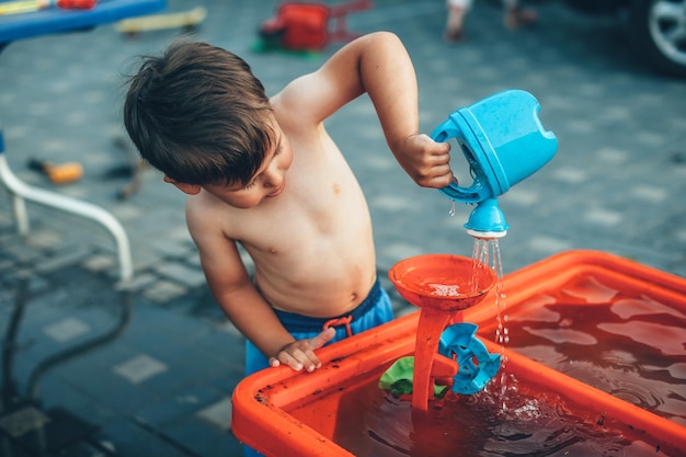 Niño caucásico sin ropa está jugando con juguetes de agua de plástico en el patio durante un día de verano