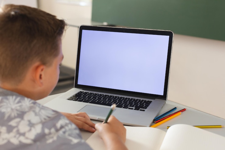 Foto niño caucásico sentado en un escritorio en el aula escribiendo y usando una computadora portátil, con espacio para copiar en la pantalla