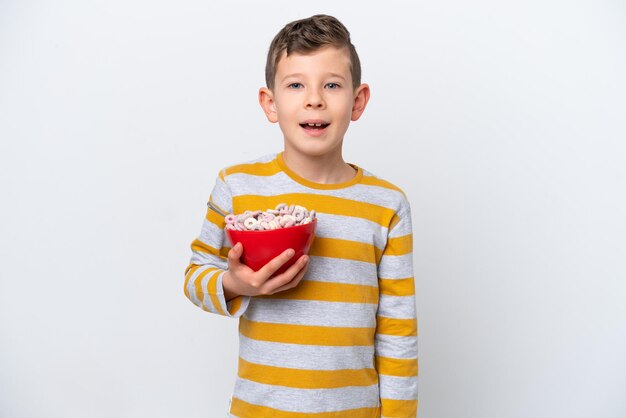 Niño caucásico sosteniendo un tazón de cereal aislado en fondo blanco con expresión facial sorpresa