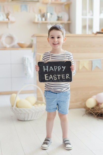 Un niño caucásico sostiene una pizarra con un cartel de Pascua en un interior de cocina claro con decoraciones de Pascua