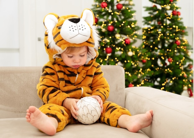 Niño caucásico con traje de tigre, símbolo del nuevo año 2022 jugando en el sofá cerca del árbol de navidad